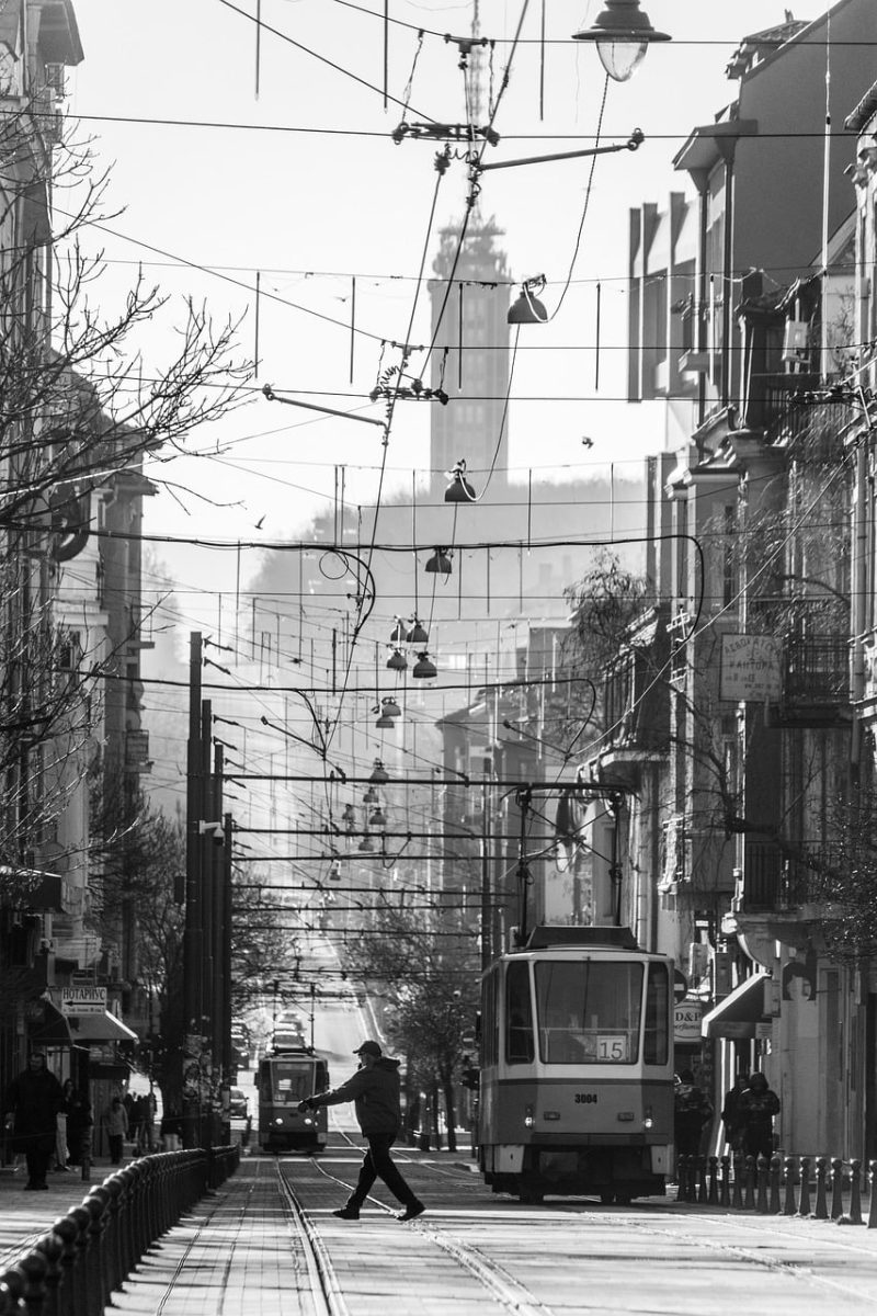 urban scene, tram, black and white photo, pedestrian, street lamp, overhead wire, winter, trees, urban transport, road, central european city, architecture, intersection, sunny day, historical tram, narrow street, building facade, traffic signal, city culture
