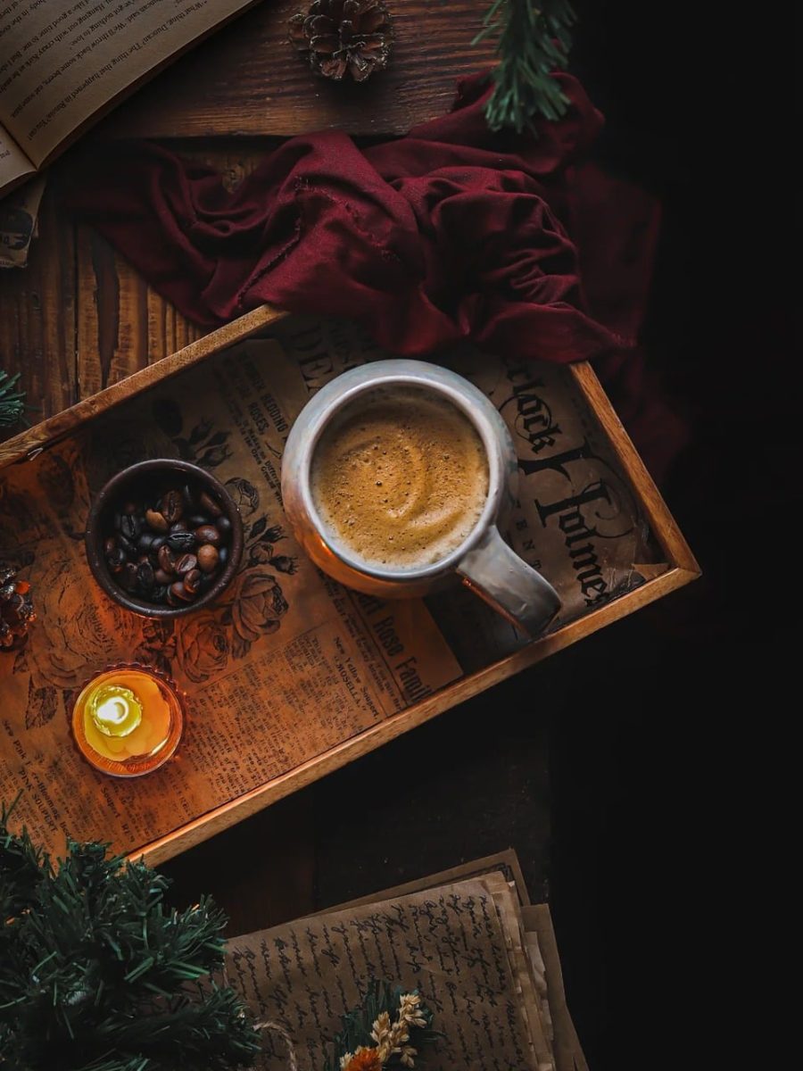 coffee mug, wooden tray, foam art, coffee beans, small bowl, lit candle, candle flame, vintage newspaper, red fabric, pinecone, moody lighting, warm aesthetic, top view, cozy morning, rustic table, caffeine, relaxation, background textures, dark food photography, green branch