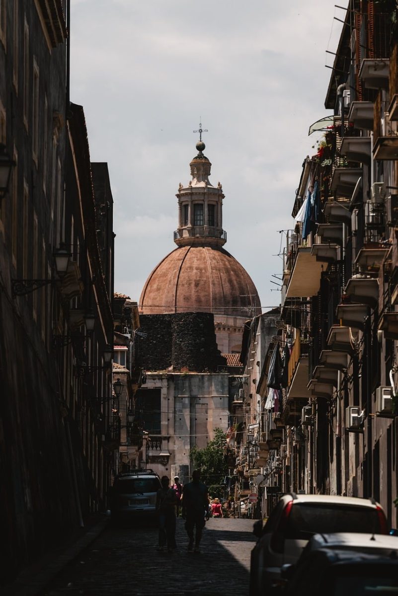 catania, cathedral, city landmark, narrow street, historic architecture, overcast sky, italian cityscape, cobblestone, pedestrians, old buildings, southern italy, travel photography, tourism, urban silhouette, city life, dome, streetscape, local architecture, culture, europe, summer day