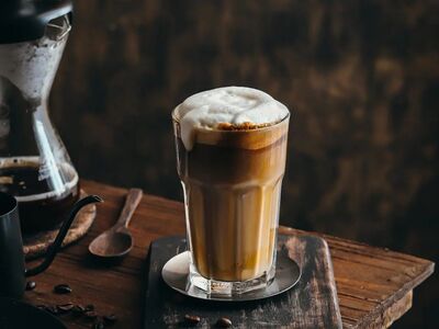 iced latte macchiato, layered espresso, milk foam, wooden spoon, chemex, coffee beans, dark textured table, metal saucer, wooden tabletop, rustic coffee maker, artistic drink, slow living, cozy morning, coffee shadow effect, warm light, wood texture, shadow play, afternoon coffee time, aesthetic photography, barista creation